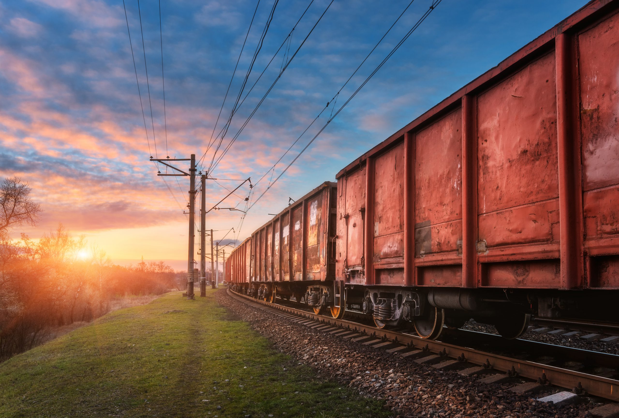 Railway station with cargo wagons and train against sunny sky with clouds in the evening. Colorful industrial landscape. Railroad with blue sky. Railway platform. Heavy industry. Cargo shipping